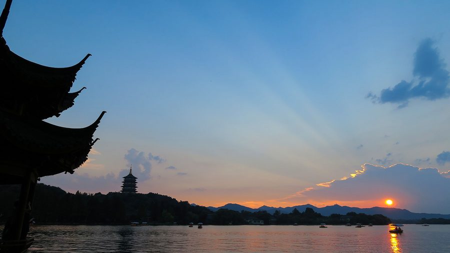 Sunset over West Lake in Hangzhou, with the silhouette of Leifeng Pagoda and a pavilion roof in the foreground.